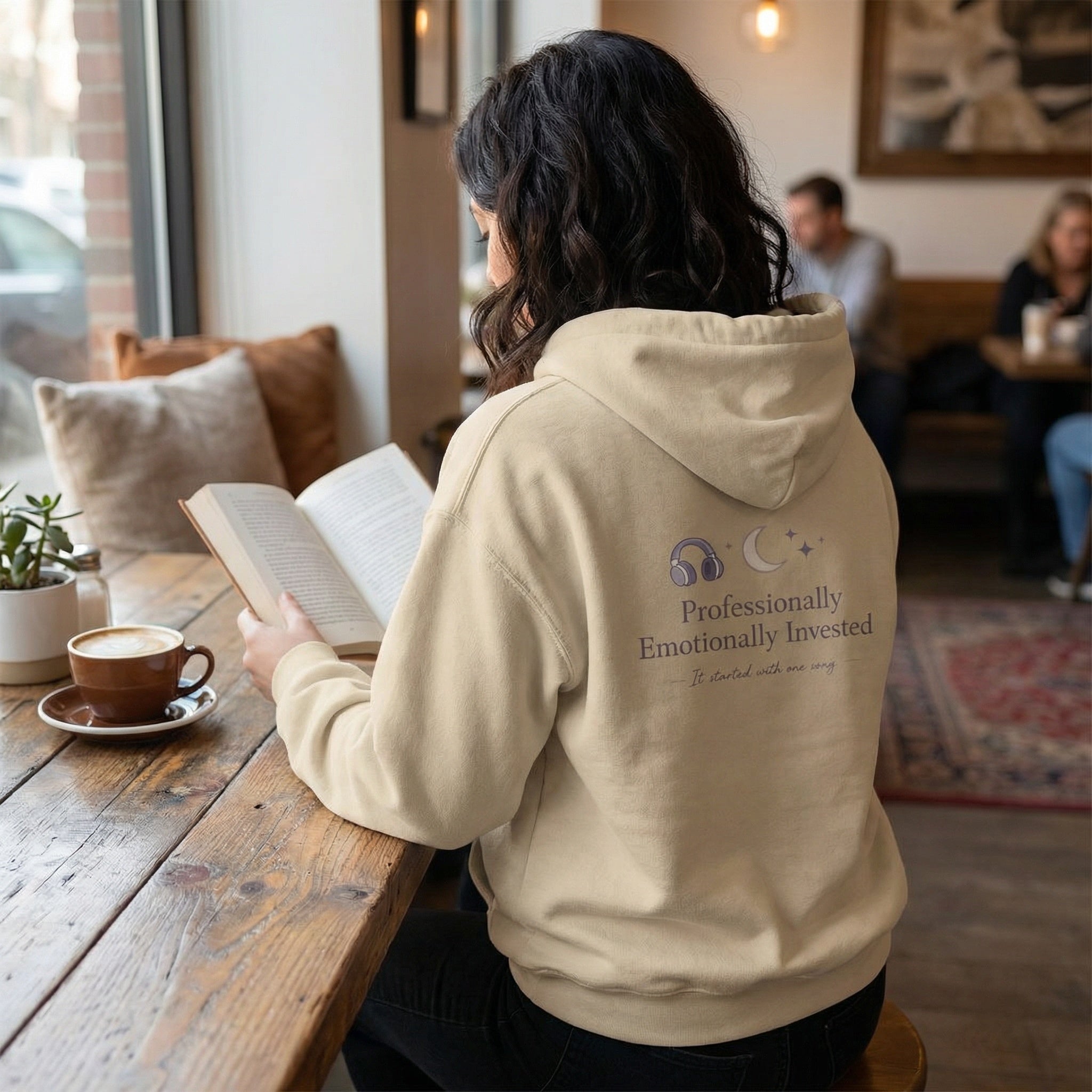 Person wearing a beige hoodie with text, sitting at a table in a cozy cafe reading a book.