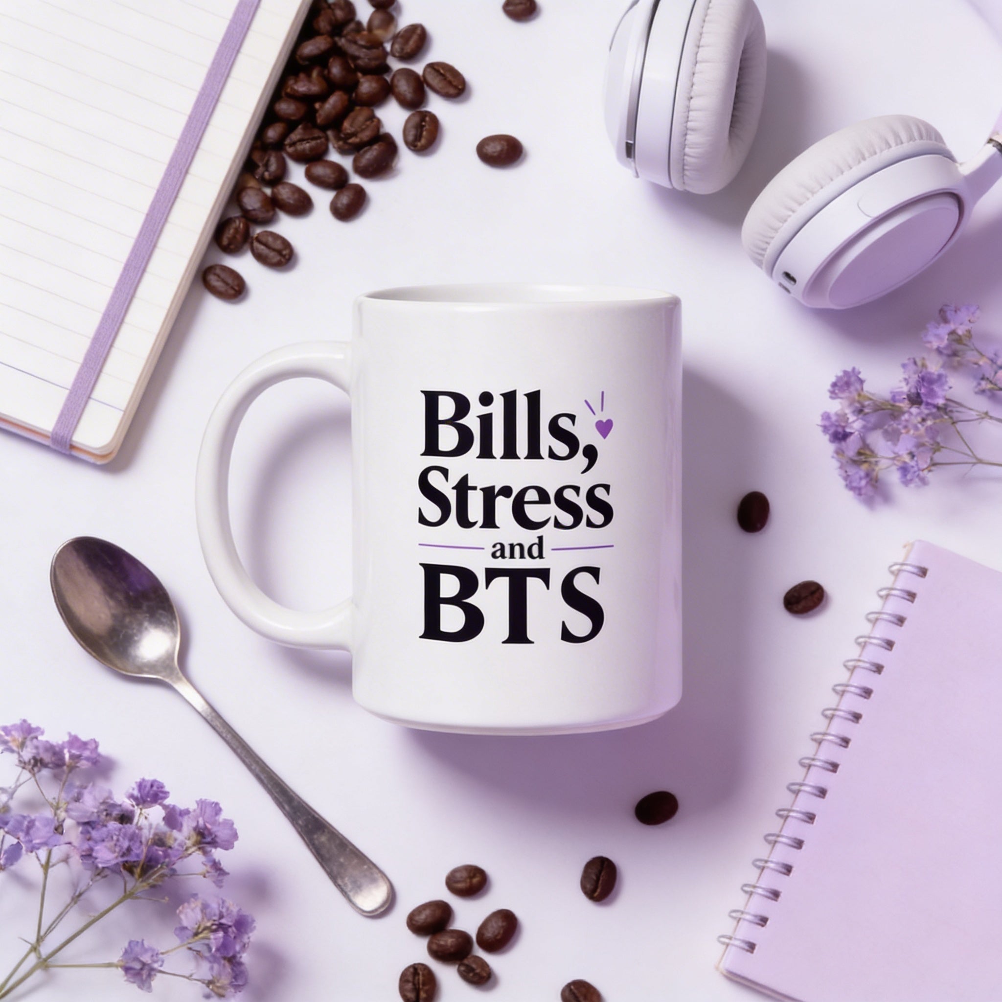 White mug with 'Bills, Stress and BTS' text on a white surface with coffee beans, headphones, and flowers.