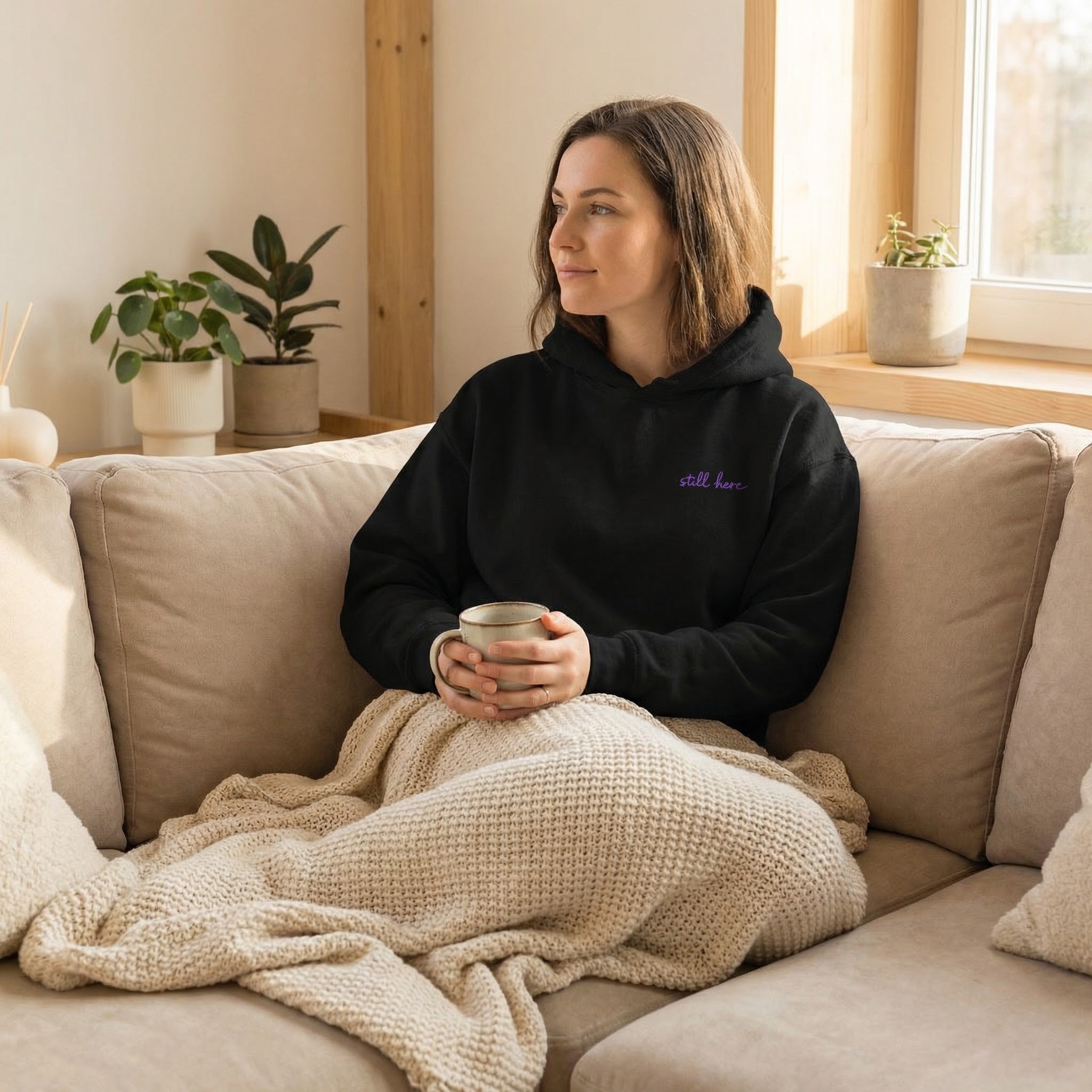 Woman sitting on a couch with a blanket and mug, wearing a black hoodie with text.