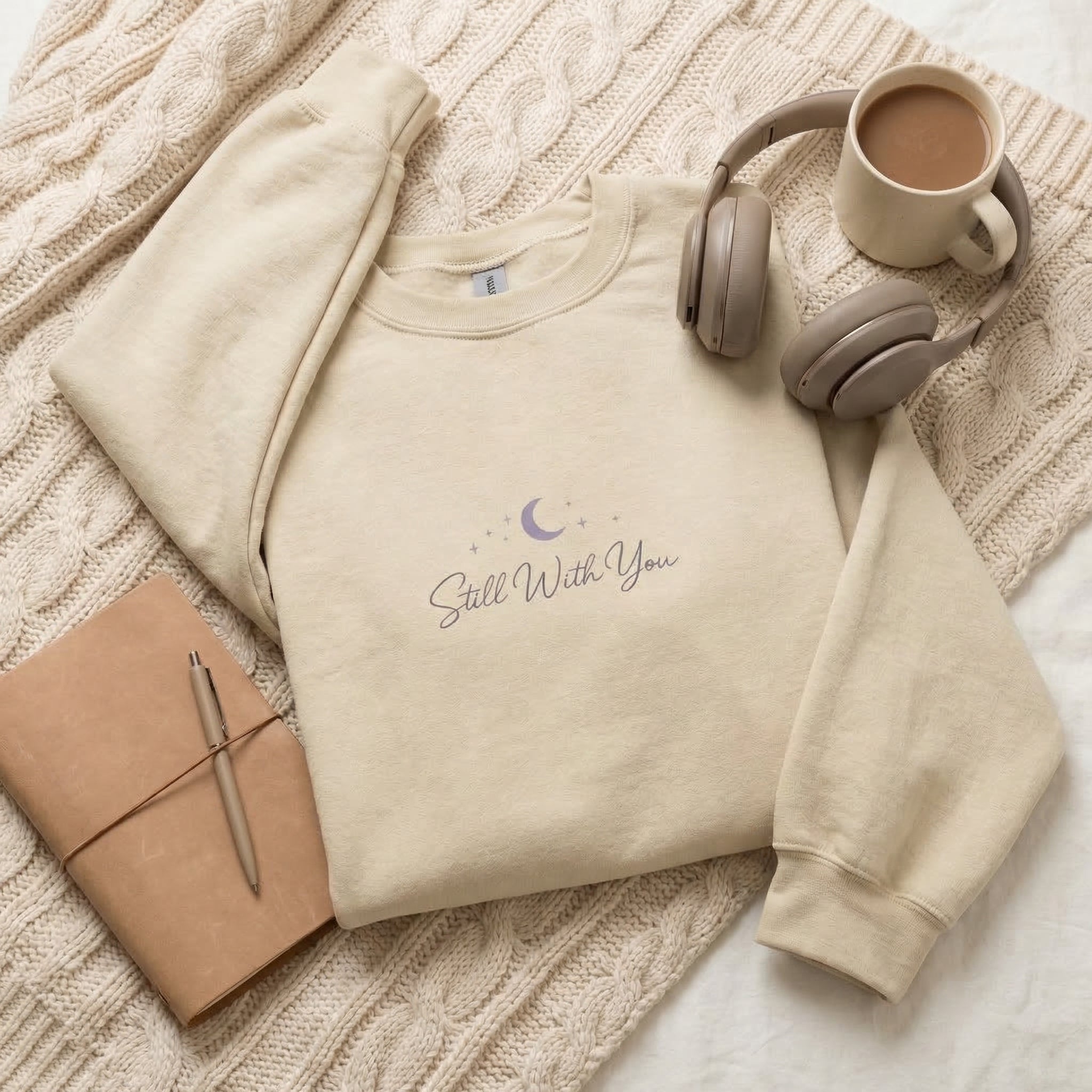 Beige sweatshirt with 'Still With You' text, coffee cup, headphones, and notebook on a textured surface