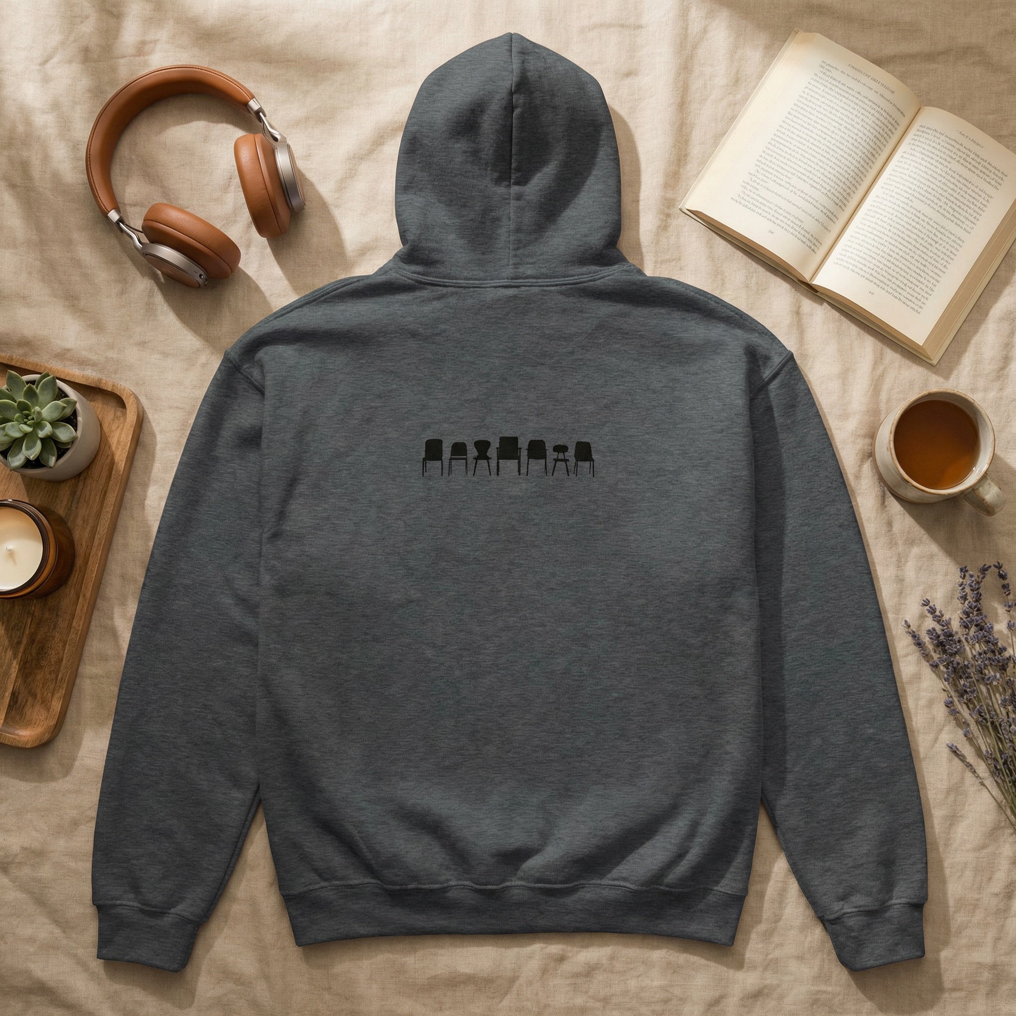 Gray hoodie with a small graphic on the back, placed on a surface with a book, cup, headphones, and plant.