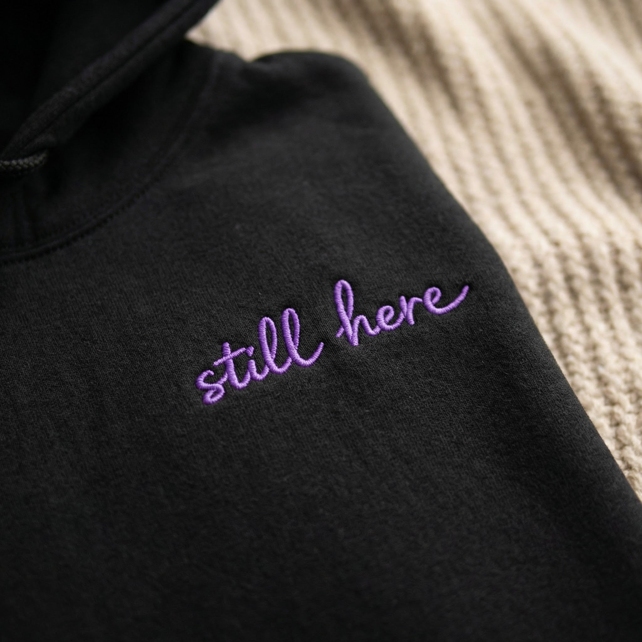 Black hoodie with 'still here' embroidered in purple on a textured surface