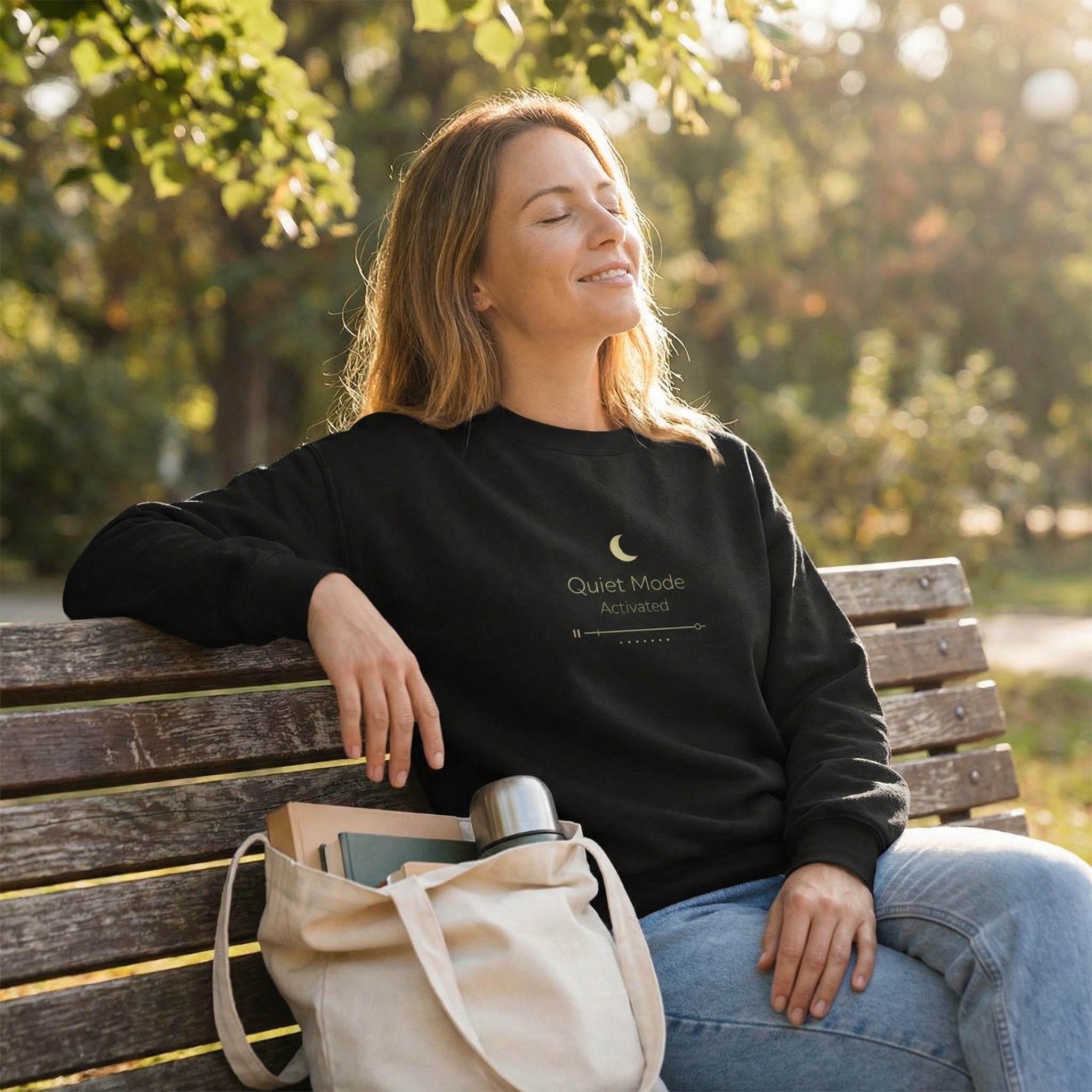 Woman sitting on a bench in a park wearing a black sweatshirt with text, holding a beige bag.
