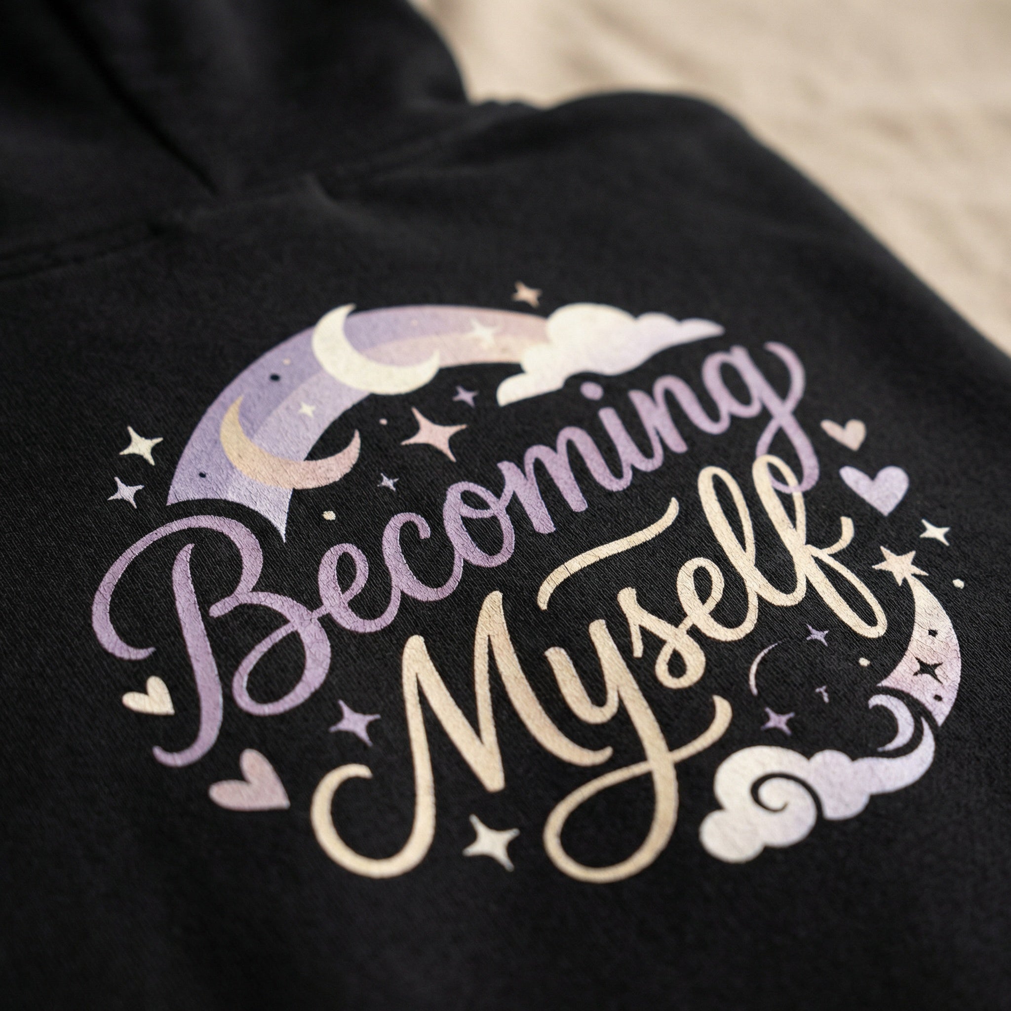 Black hoodie with 'Becoming Myself' text and celestial design on a neutral background