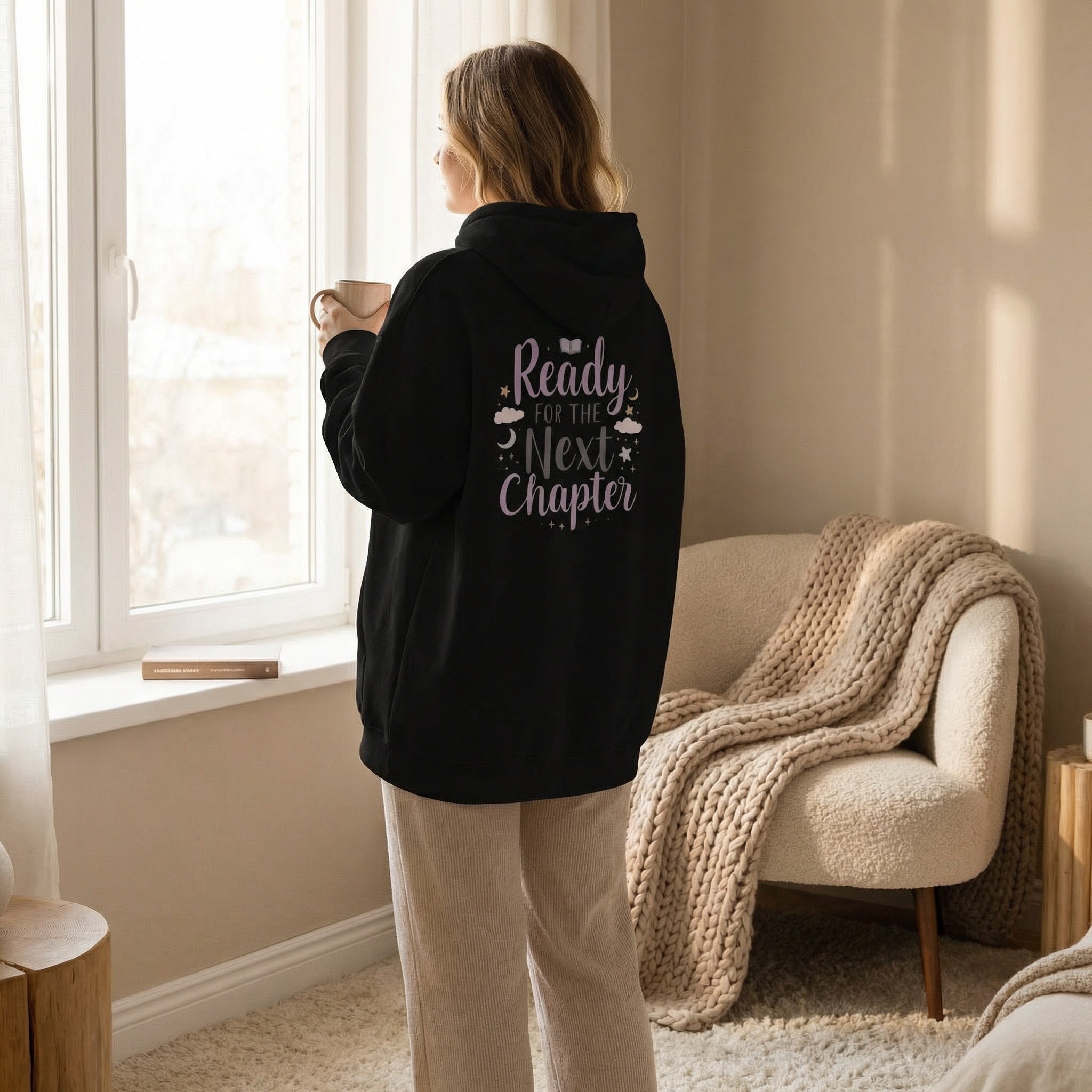 Person wearing a black hoodie reading "Ready for the Next Chapter," standing by a window holding a mug.