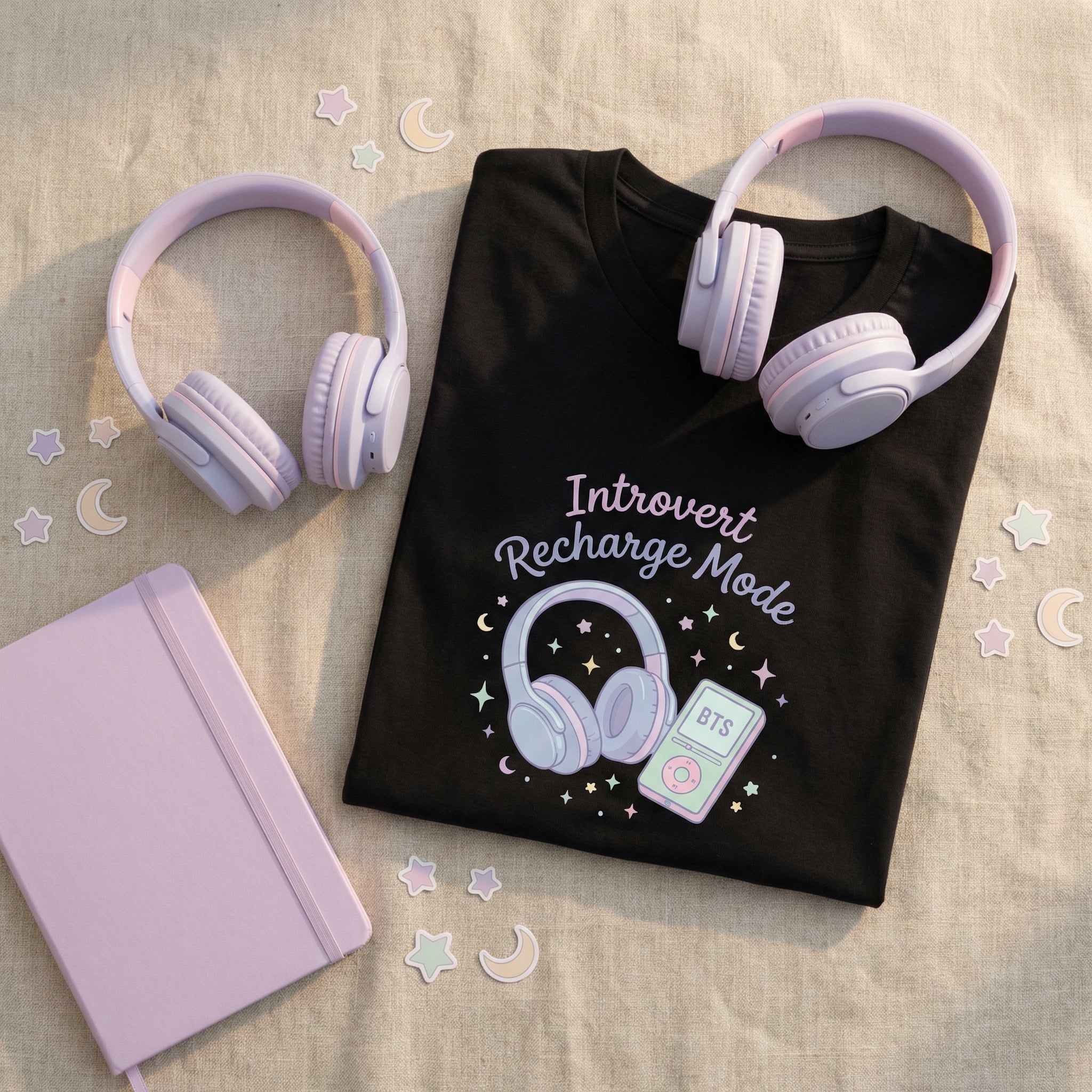 Black t-shirt with 'Introvert Recharge Mode' design, pink headphones, and pink notebook on a beige surface.