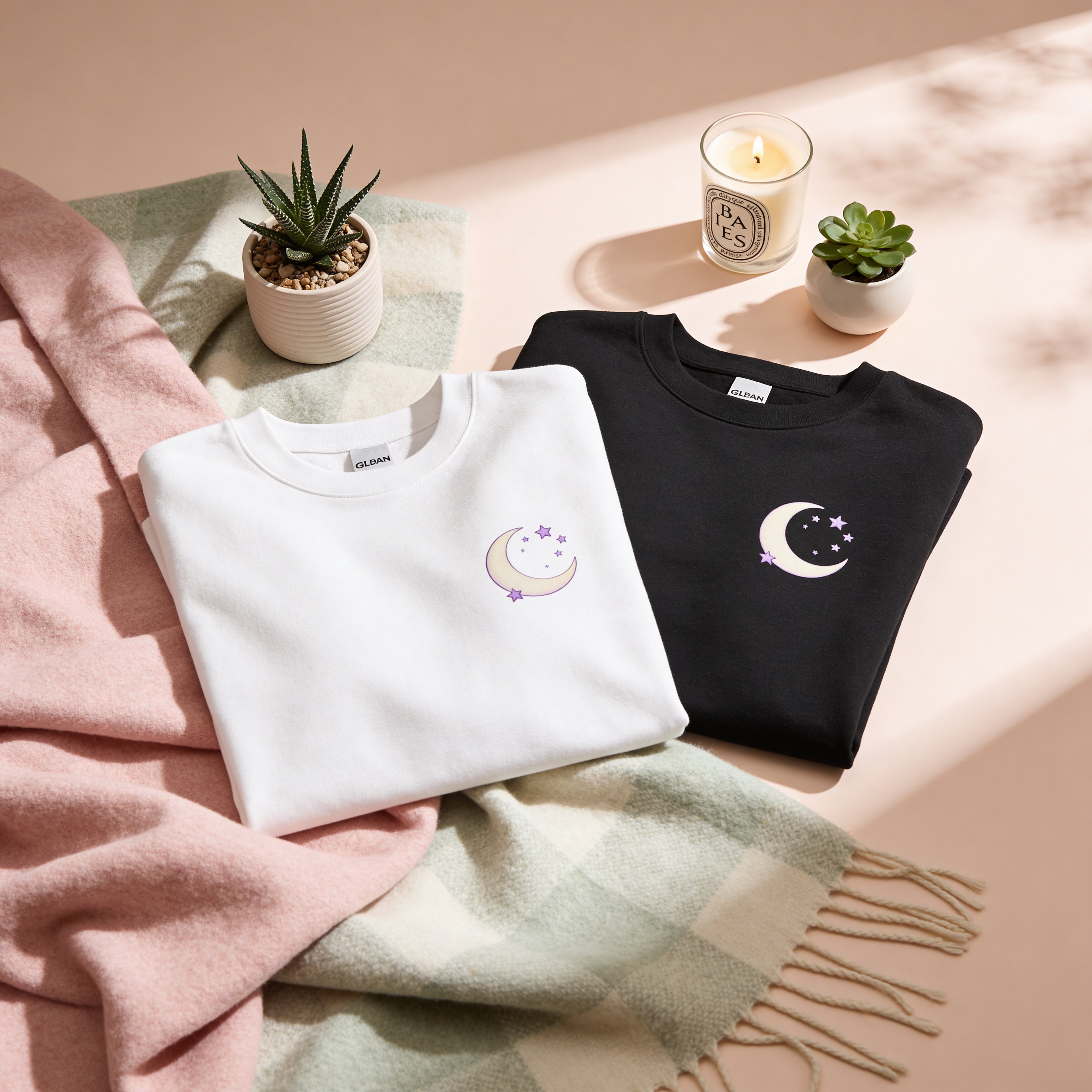 Two sweaters, one white and one black, with moon and star designs on a soft surface with plants and a candle.