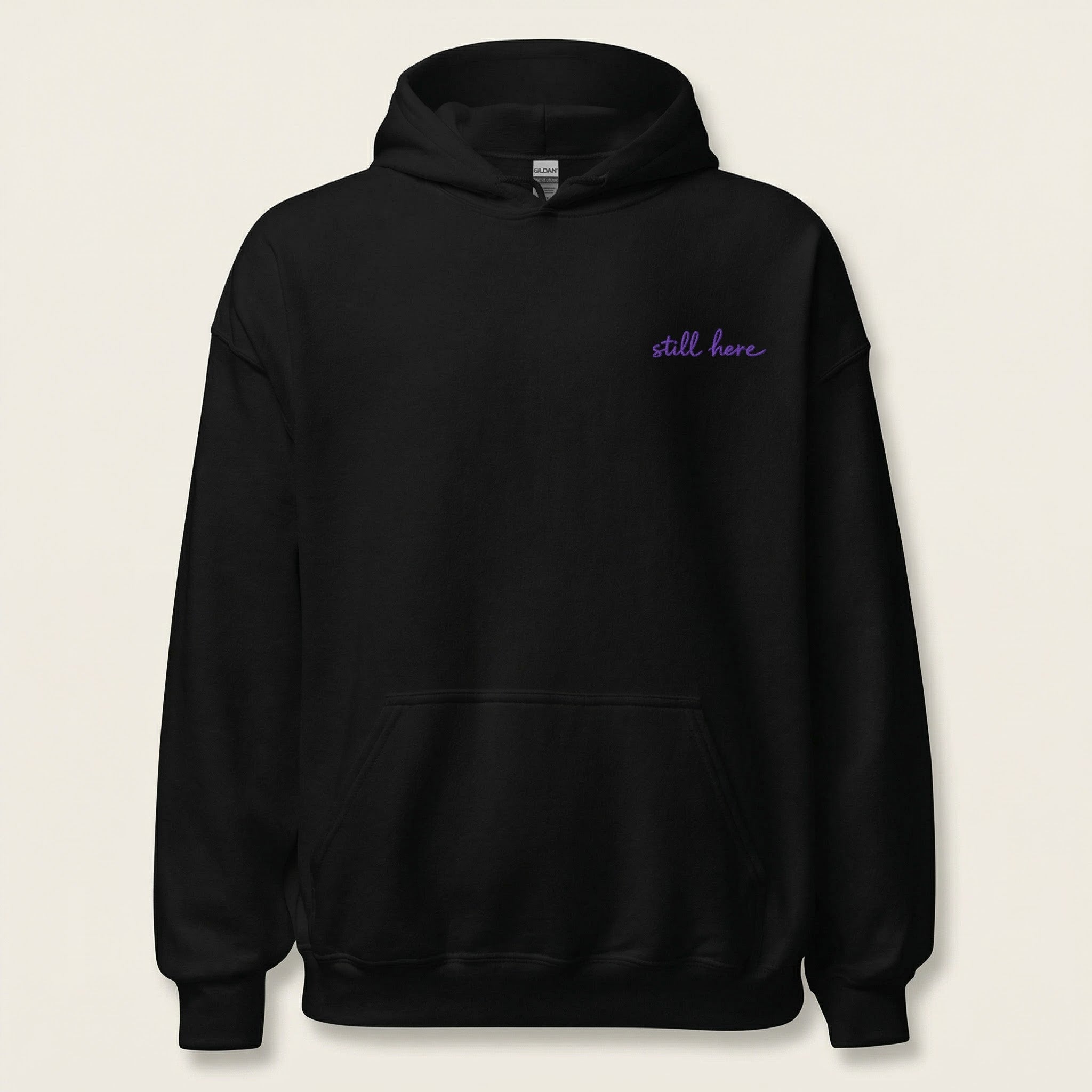 Black hoodie with 'still here' text in purple on a beige background
