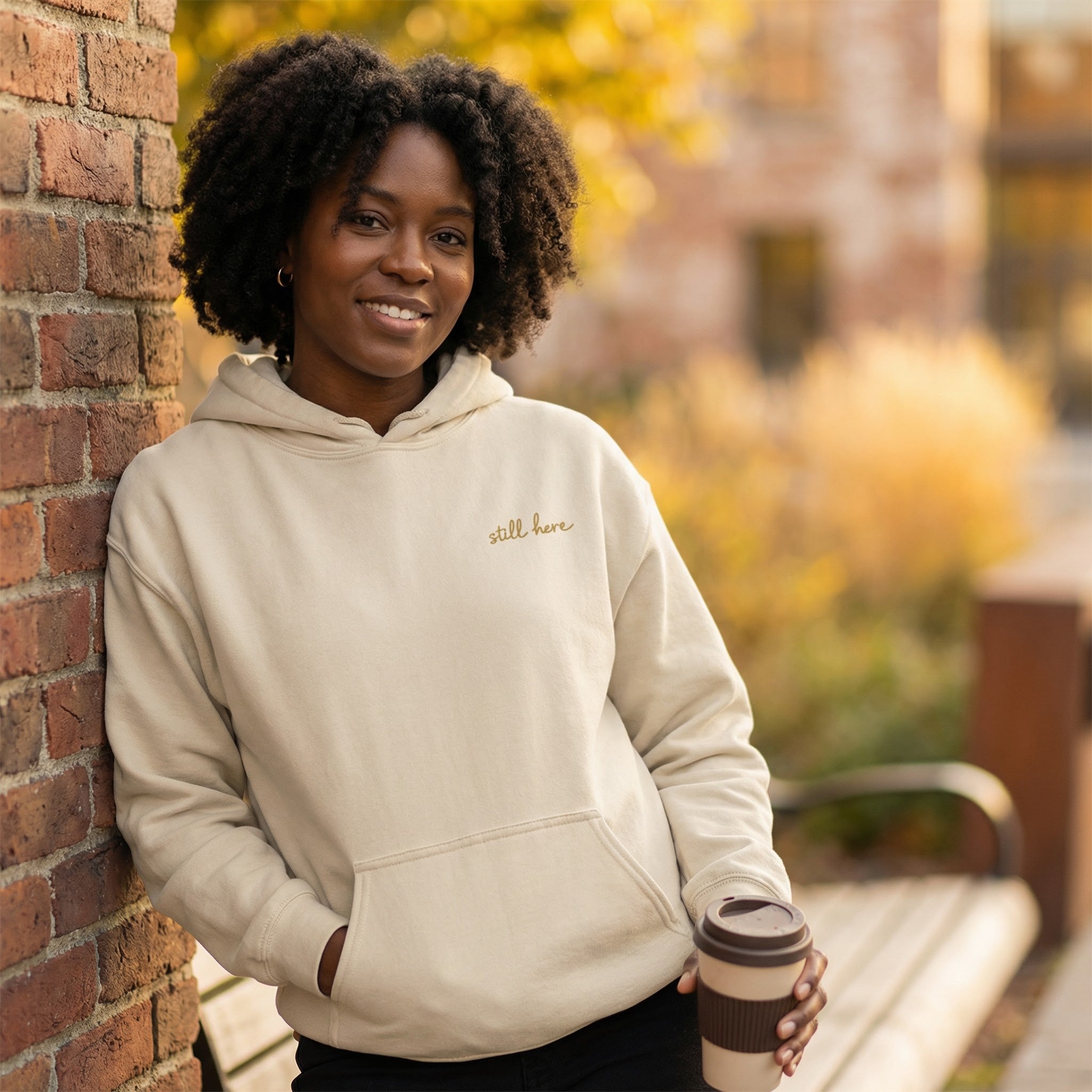 Person wearing a beige hoodie with 'still here' text, holding a coffee cup, leaning against a brick wall outdoors.